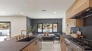 A modern kitchen with a central island, dark countertops, wooden cabinets, and a large window providing a view of the outdoors. - The Dona Modern (Studio City)
