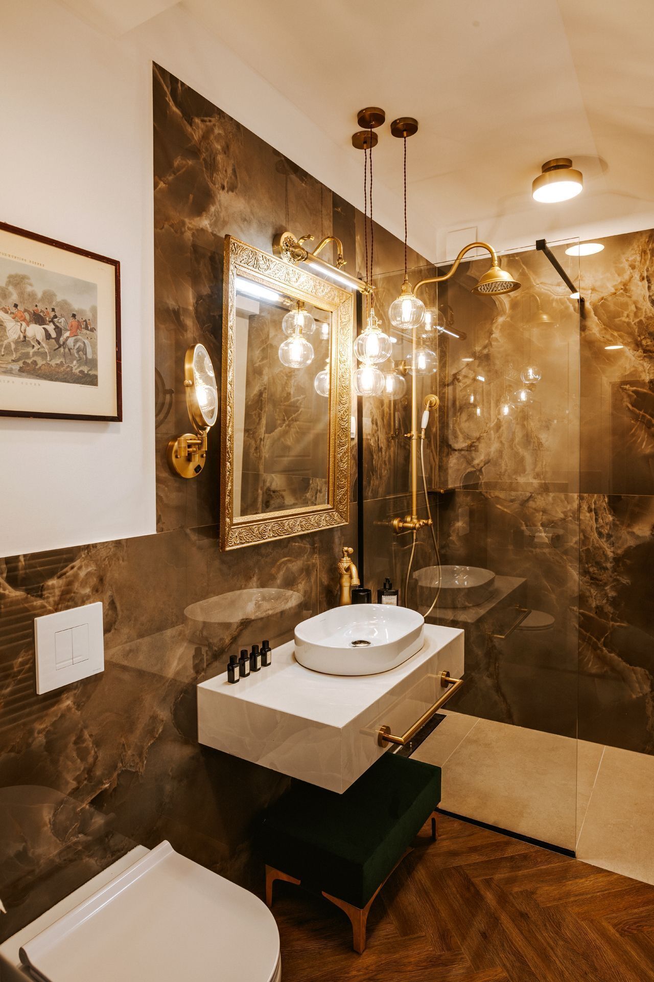 Ein Badezimmer mit Marmorwand, einem großen Spiegel, einem spiegel mit goldener Rahmung, einem weißen Waschbecken, einer Dusche und einem Holzboden.