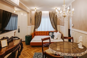 Un dormitorio con cama, una mesa redonda con mantel de encaje, una silla de madera, un reposapiés verde, una araña y un televisor.