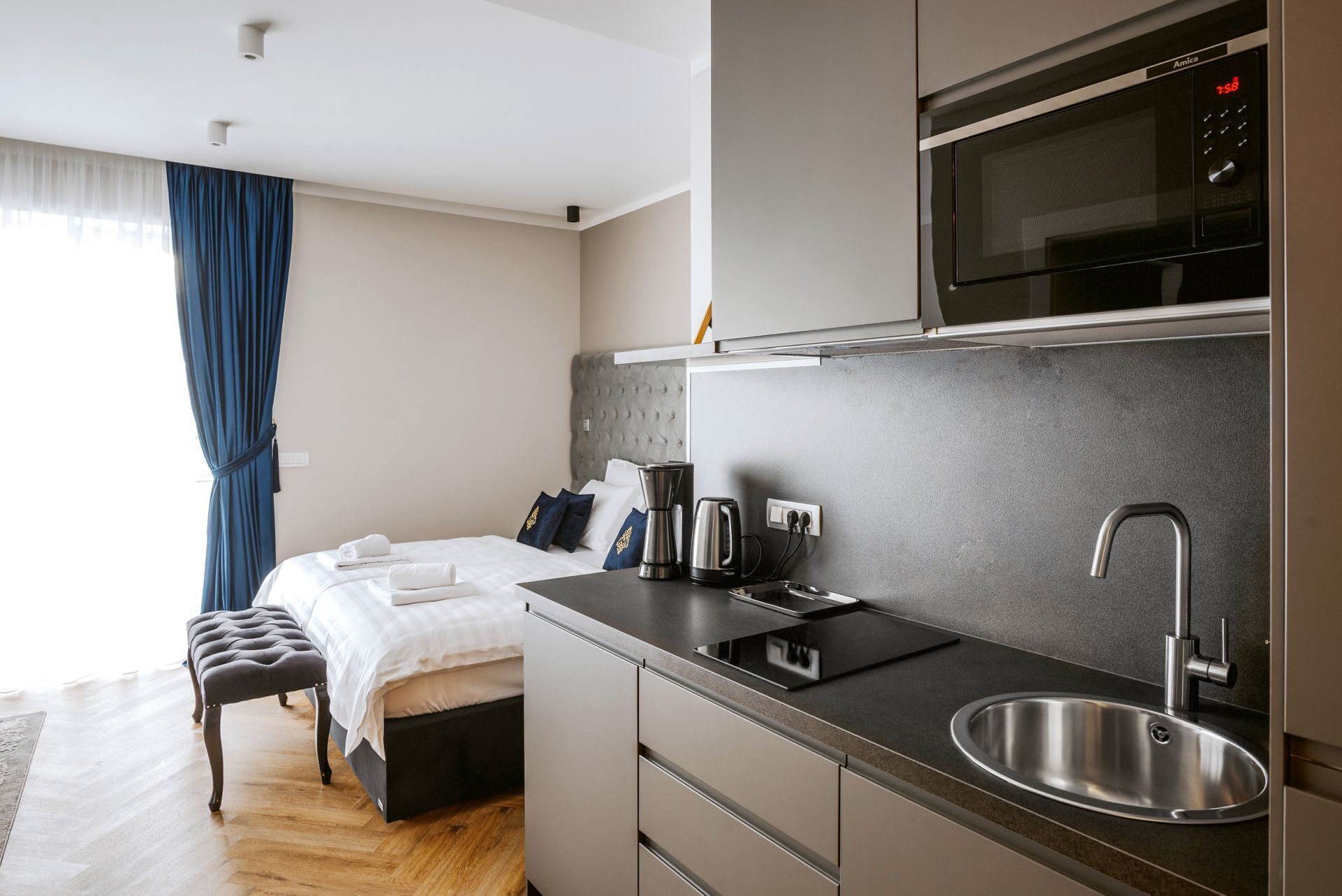 Una habitación de hotel moderna con una cama, una kitchenette equipada con microondas, un lavamanos y una mesita de noche con cafetera.