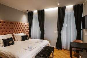 Quarto de hotel com cabeceira estofada, cama com roupa de cama branca, um banquinho preto, uma escrivaninha e cortinas leves.