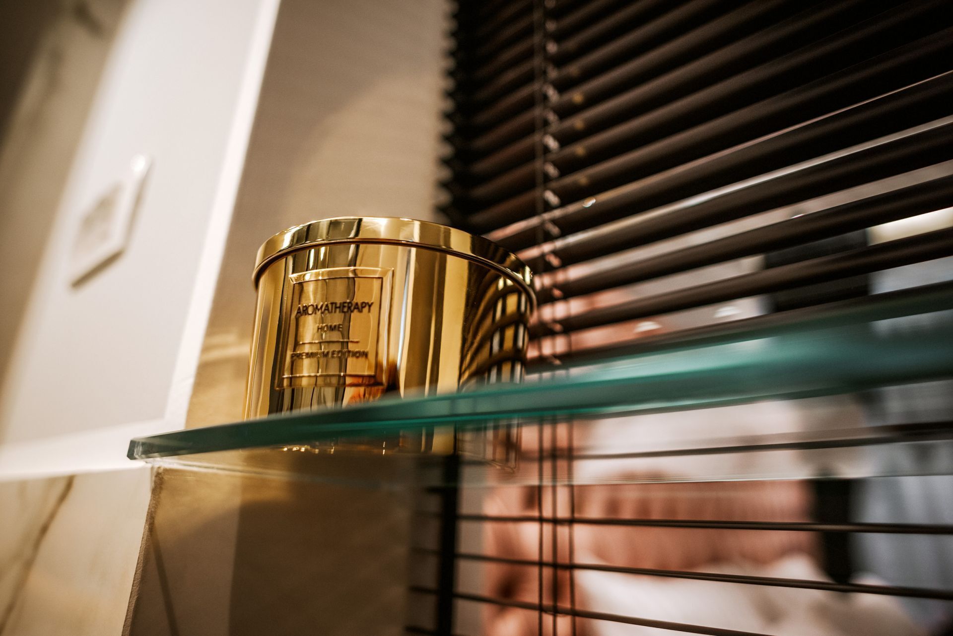 Un recipiente dorado etiquetado 'AROMATHERAPY HOME PREMIUM EDITION' sobre una estantería de vidrio.