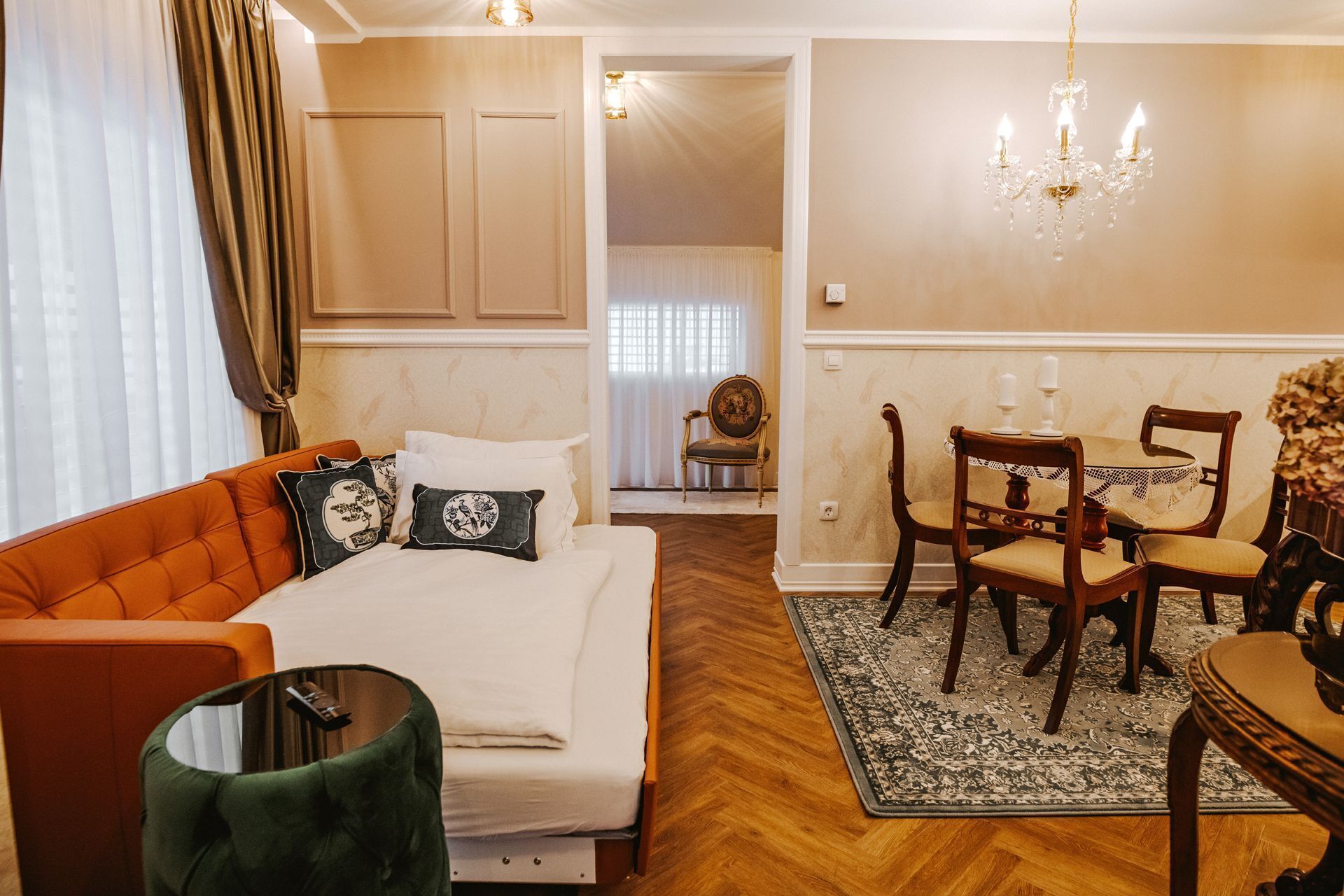 A hotel room with a bed, a sofa, a dining table with chairs, a chandelier, and a view of another room.