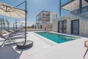 Ein moderner Poolbereich mit Liegestühlen und Sonnenschirmen, eine Steinmauer und ein gläserner Gebäudebereich.