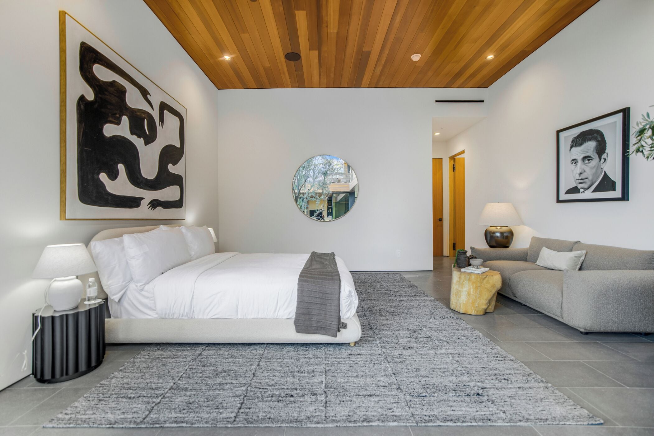 A modern bedroom with a bed, a sofa, a round mirror, and artwork on the walls.