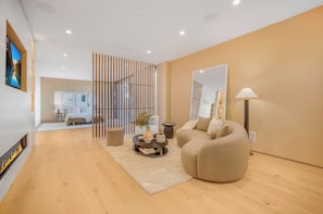 A modern living room with a beige sofa, a round coffee table with decorative items, a fireplace, and a large mirror on the wall. - Oak Pass Manor (Beverly Hills)