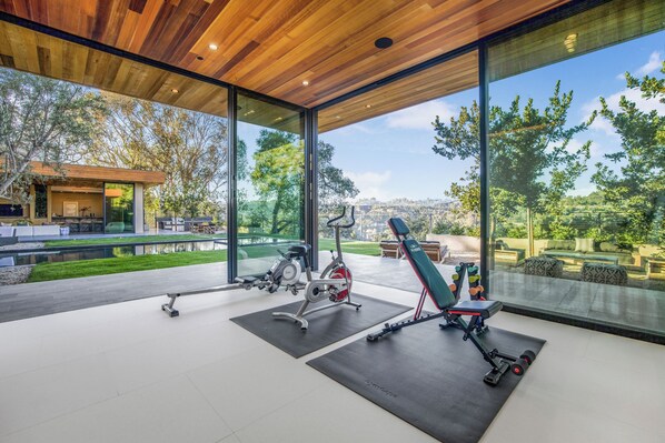 A modern gym with exercise equipment, large windows, and a view of a landscaped garden. - Oak Pass Manor (Beverly Hills)