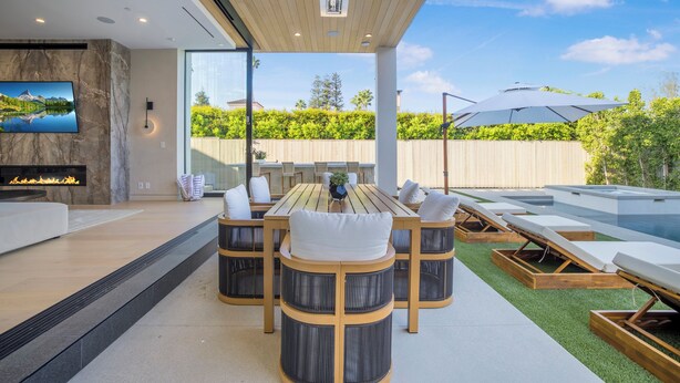Un espacio moderno para comer al aire libre con mesa y sillas de madera, un televisor montado en la pared y vistas al jardín trasero.