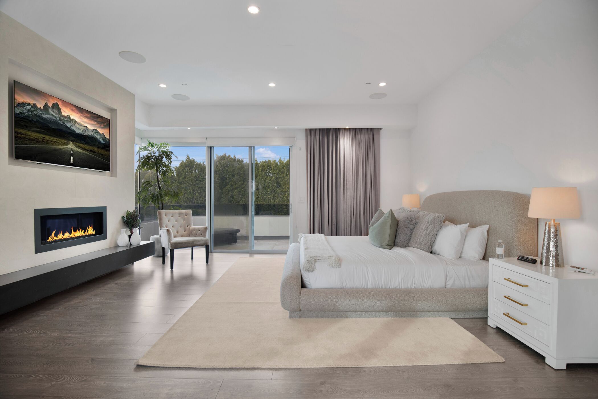 A modern bedroom with a large bed, a fireplace, a view through glass doors, and a mounted TV.