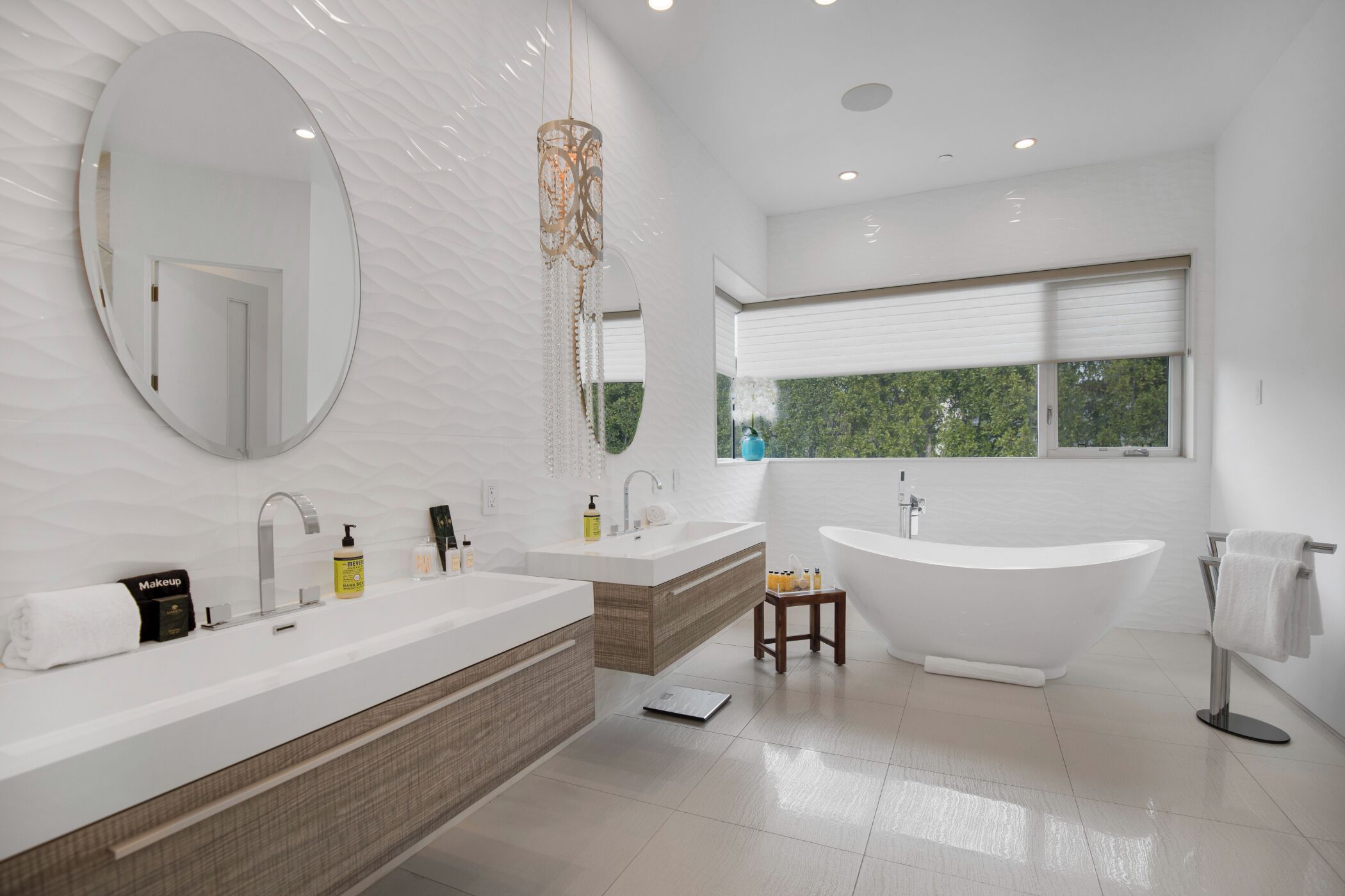 Un baño moderno con un espejo ovalado grande, dos lavamanos, una bañera independiente y una ventana con vistas a zonas verdes.
