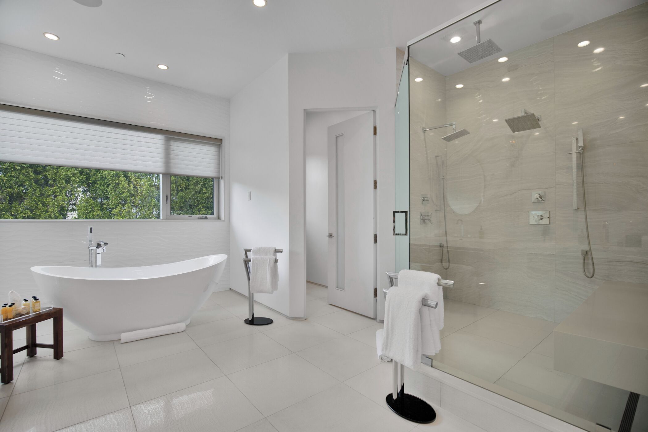 A modern bathroom with a freestanding bathtub, a glass-enclosed shower, and a large window providing a view of trees.