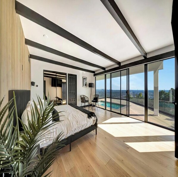A modern bedroom with a large bed, a TV, and a view of a city skyline and pool. - Skyview Estate (West Hollywood)