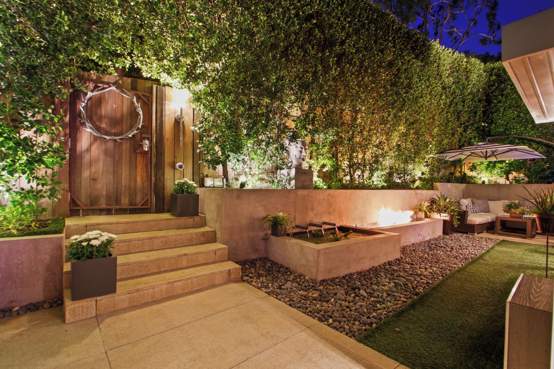 A well-lit outdoor patio area with a wooden door, steps leading to a seating area, and a fire feature.