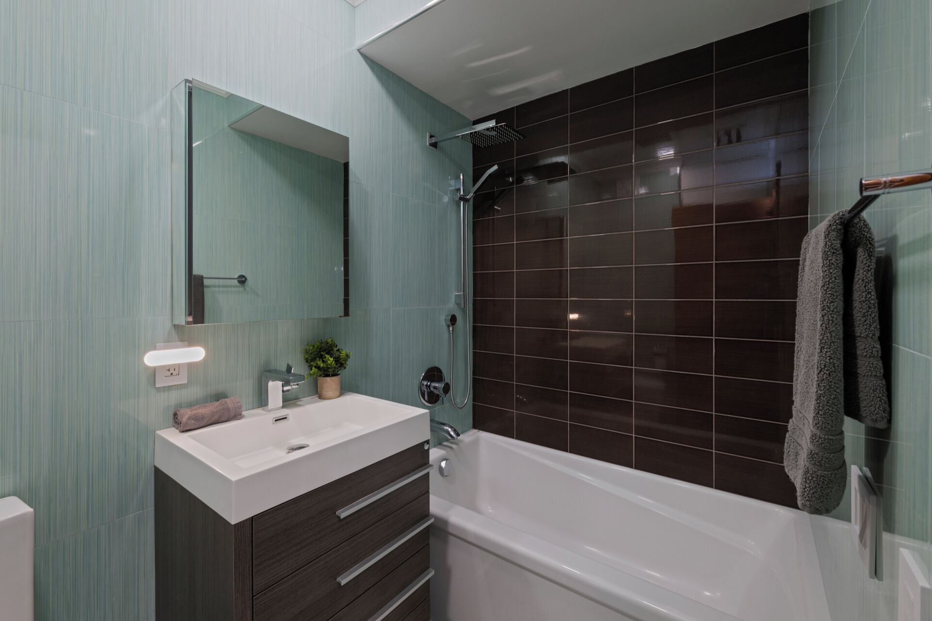 Une salle de bain moderne avec un lavabo blanc, un meuble-lavabo en bois foncé, un grand miroir et une douche carrelée aux carreaux foncés.