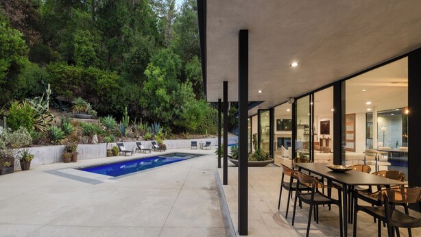 En moderne udendørs terrasse med en pool, omgivet af frodigt grønt og et spiseområde med moderne møbler.