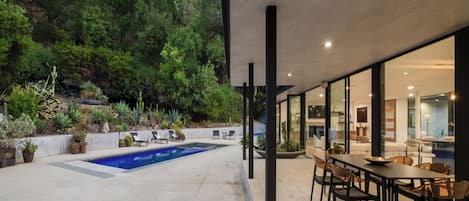 Un patio exterior moderno con piscina, rodeado de abundante vegetación y un área de comedor con mobiliario contemporáneo.