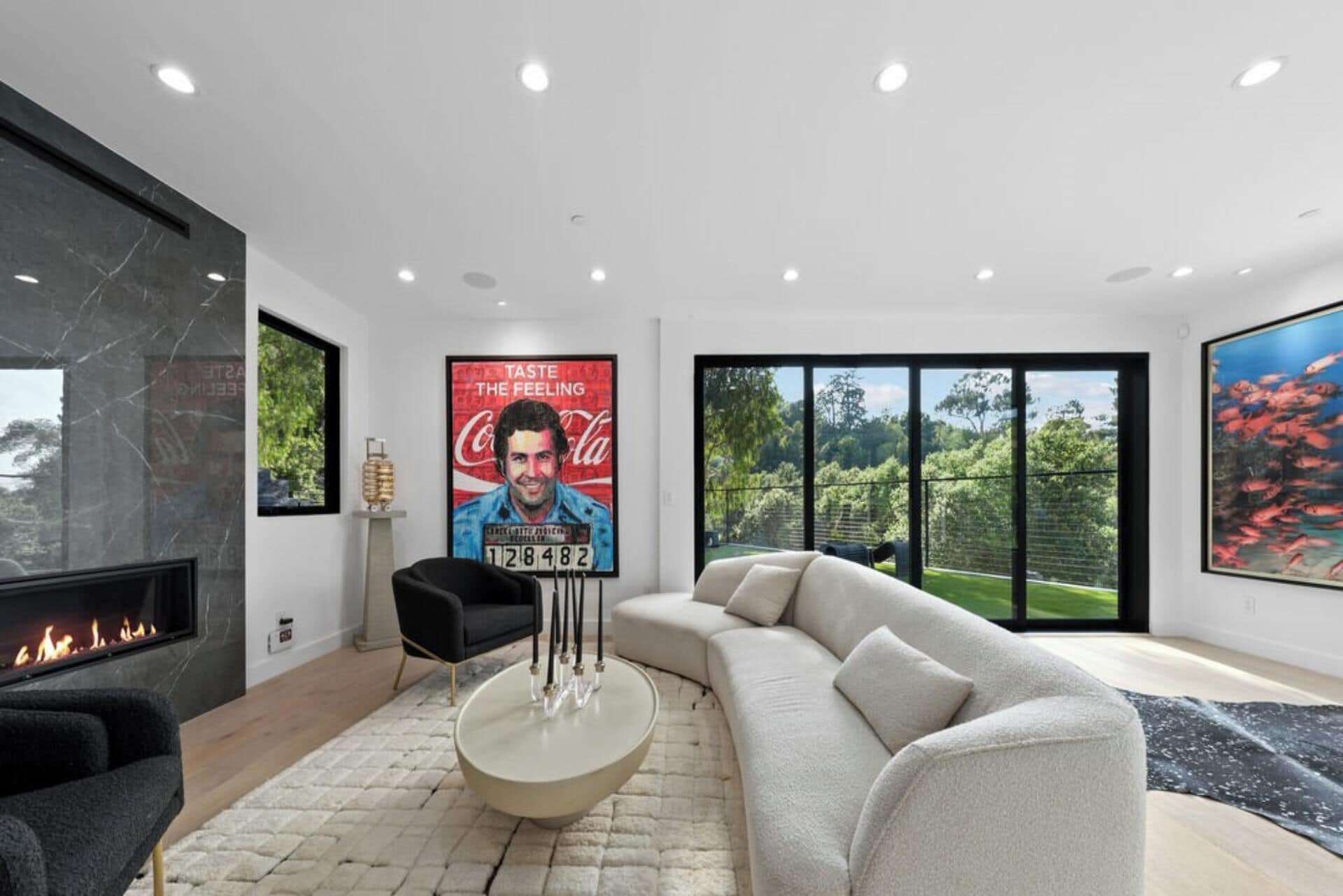 Uma sala de estar moderna com lareira, um sofá grande e uma mesa de centro. Há um pôster na parede e uma janela com vista para o verde.