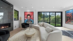 A modern living room with a fireplace, a large sofa, and a coffee table. There is a poster on the wall and a window with a view of greenery. - Oak Haven Sanctuary (Studio City)