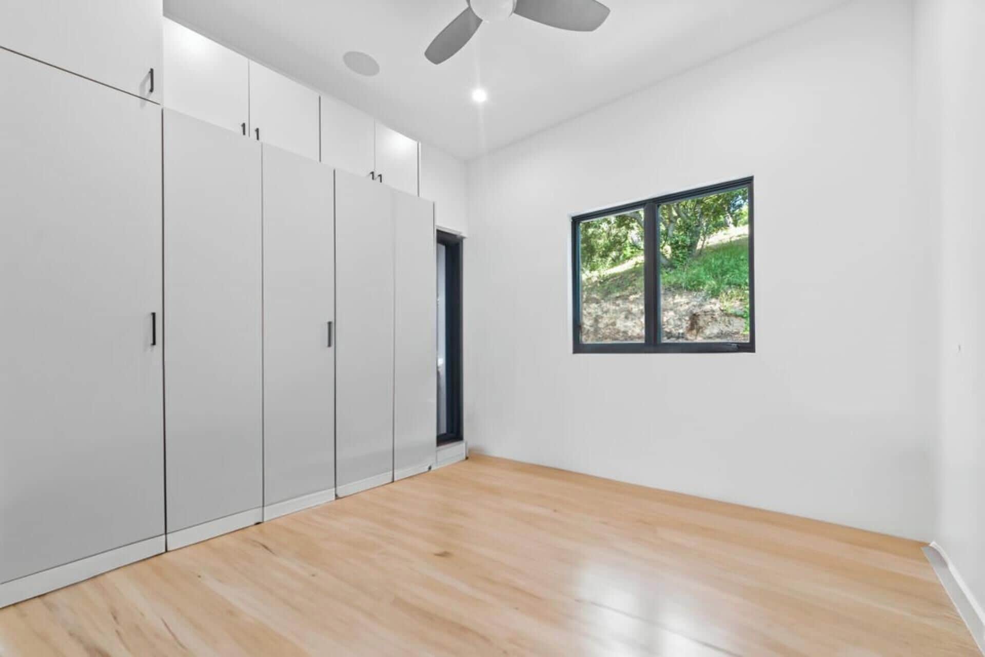 A room with a large wardrobe, a window with a view of greenery, and a ceiling fan.