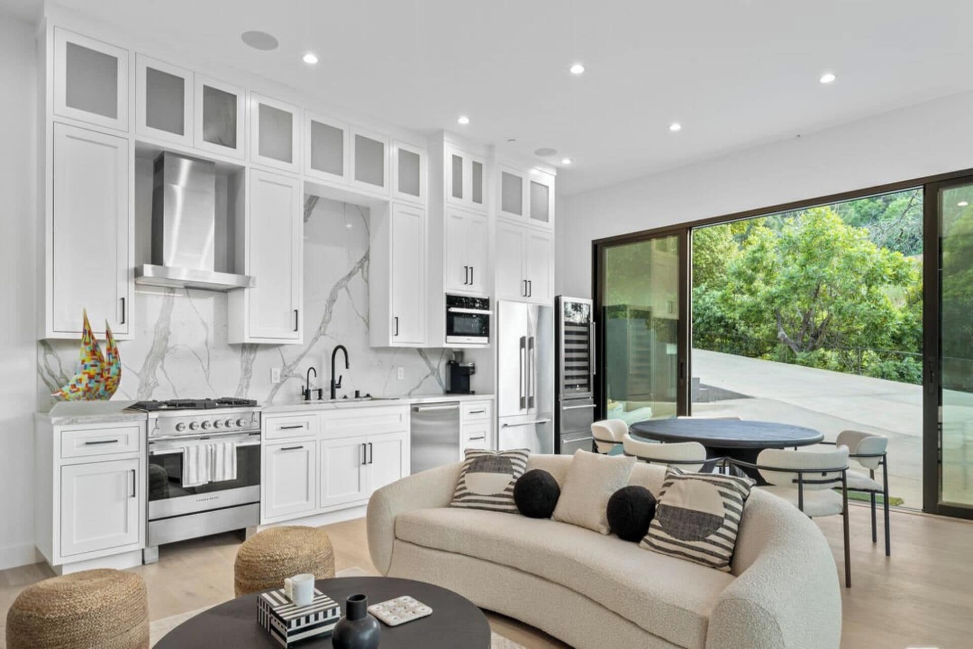 A modern kitchen with white cabinetry, a dining area with a round table and chairs, and a large sliding glass door leading to a green outdoor area.