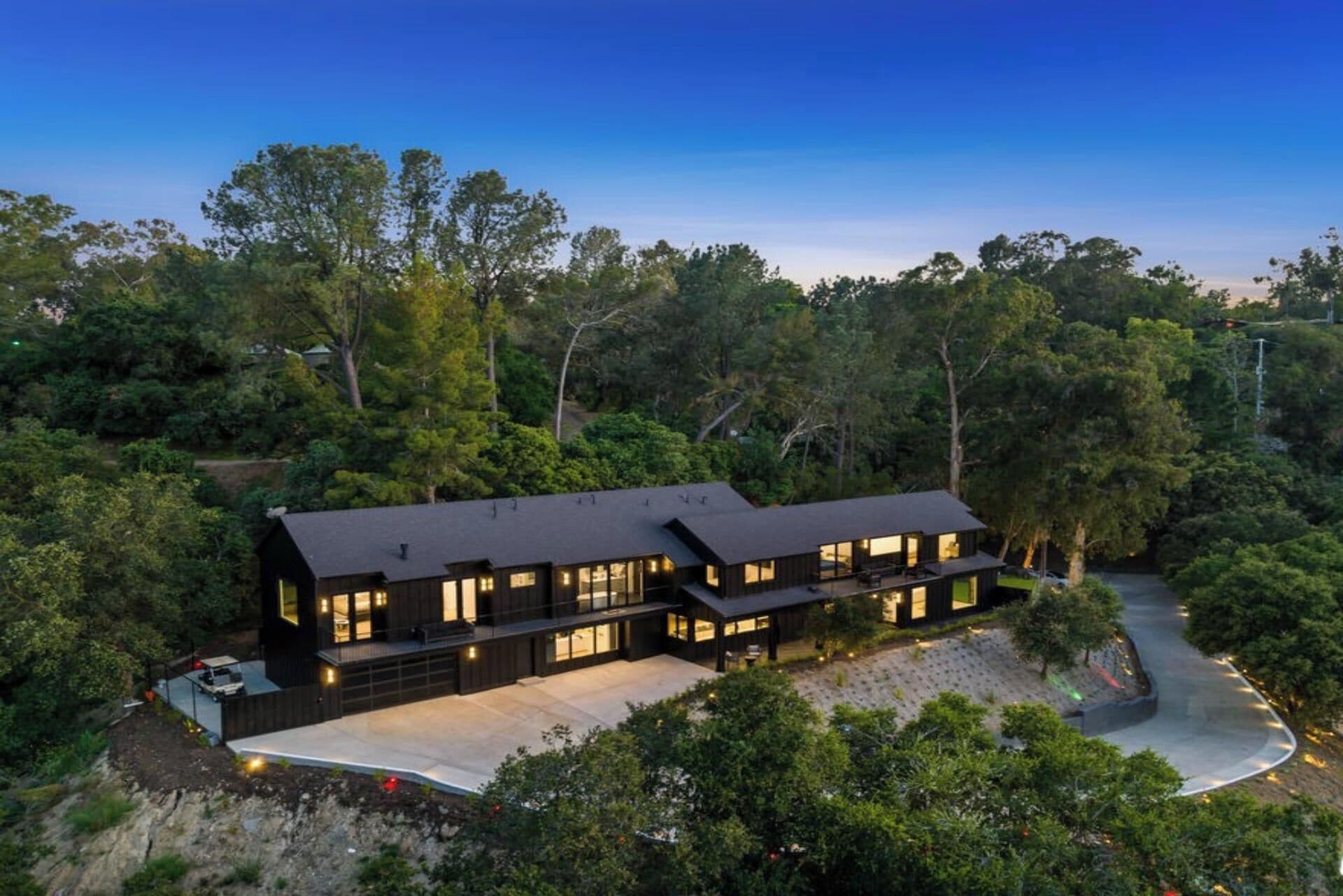 Uma casa moderna de dois andares com telhado escuro, rodeada por árvores densas.