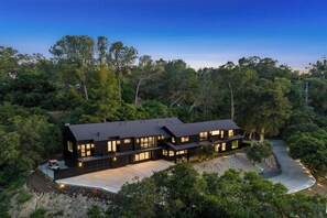 Uma casa moderna de dois andares com telhado escuro, rodeada por árvores densas.