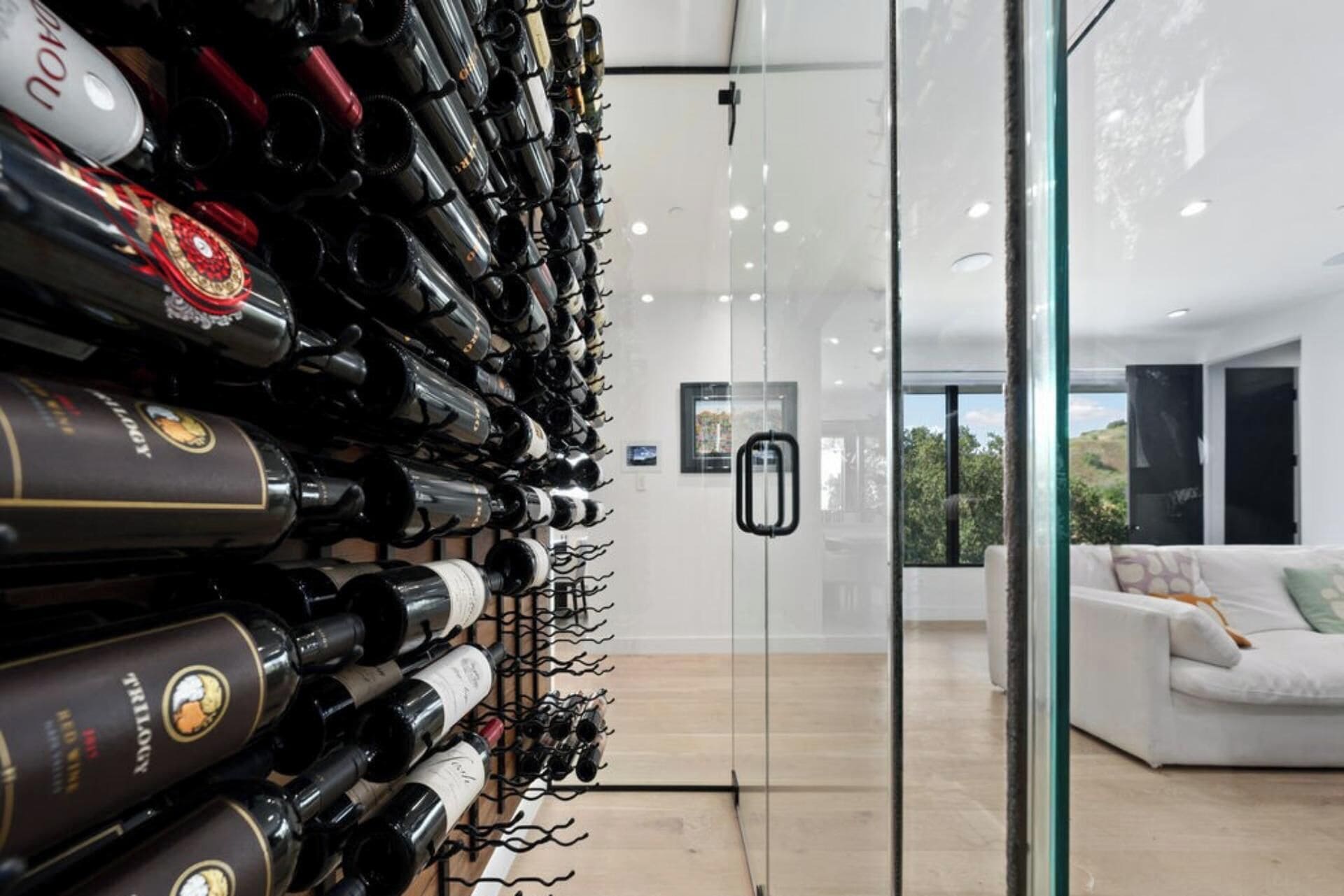 Uma adega com garrafas de vinho, uma porta de vidro que dá para um espaço com sofá branco e vista para o verde lá fora.