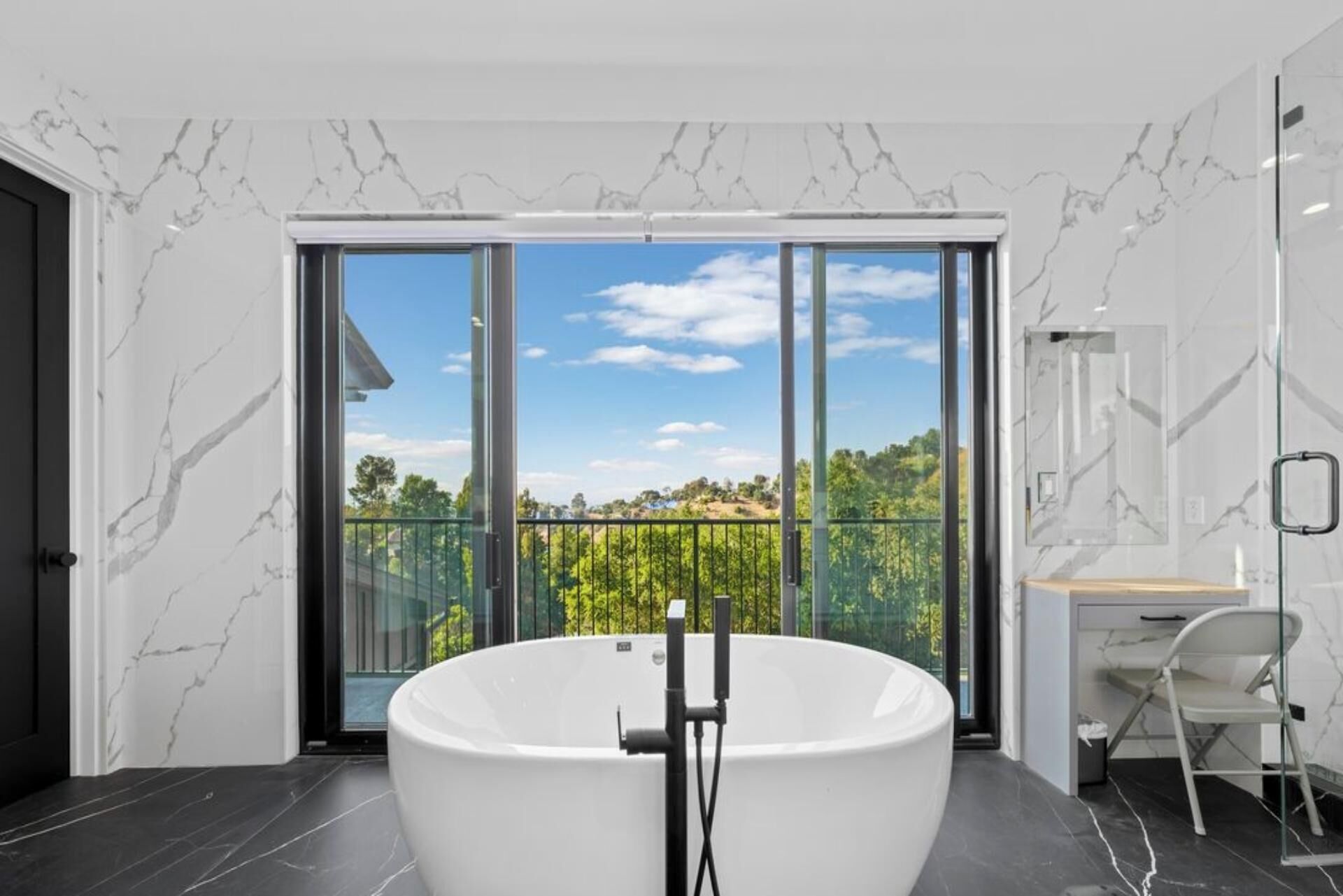 A modern bathroom with a freestanding bathtub, large sliding glass doors leading to a balcony, and a minimalist desk area.