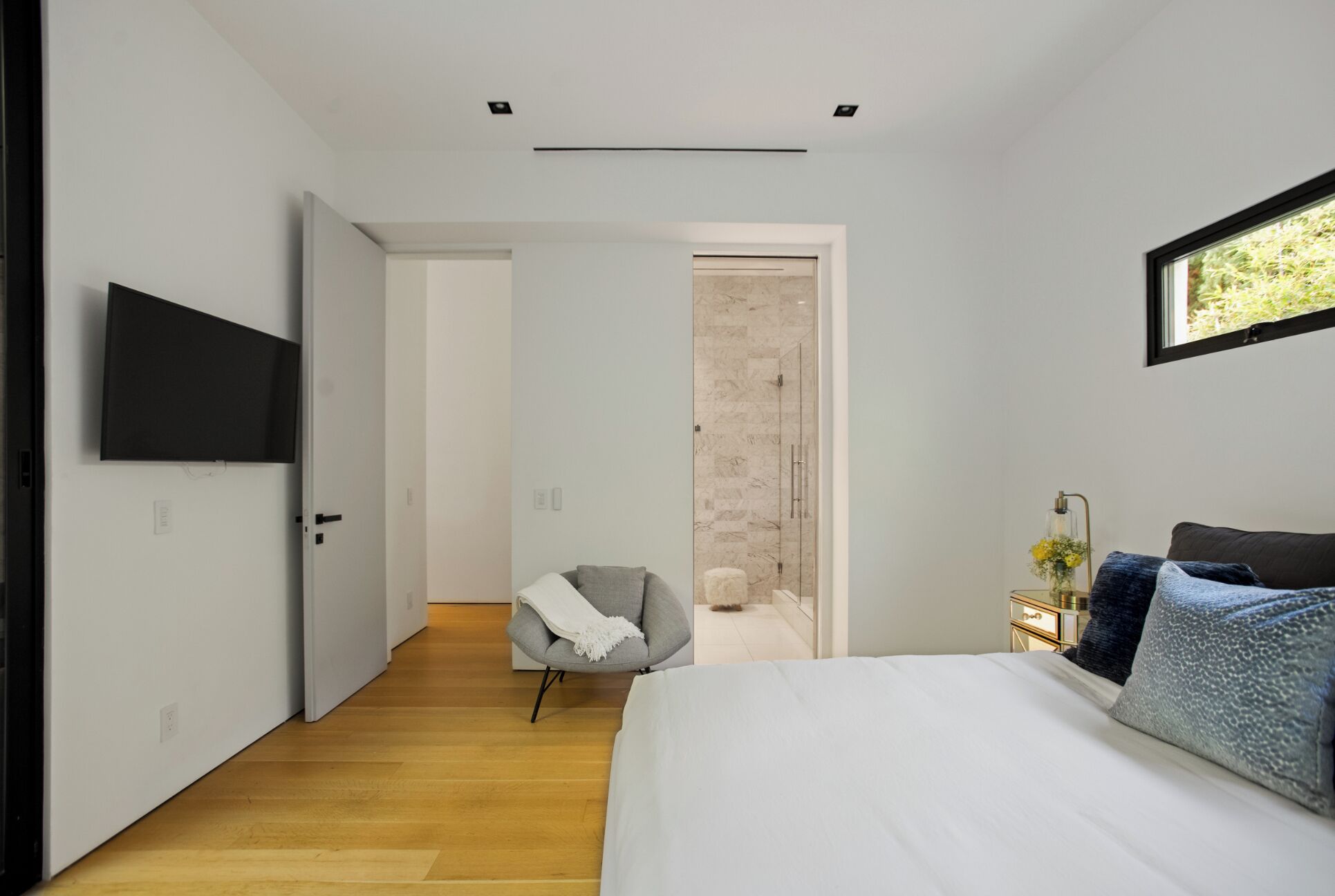Ein modernes Schlafzimmer mit einem Bett, einem an der Wand befestigten Fernseher, einem Sessel und einem kleinen Tisch mit einer Lampe.