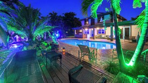 A swimming pool with a wooden deck, lounge chairs, and a house with illuminated windows. - Blockchain Manor (North Miami Beach)
