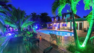 A swimming pool with a wooden deck, lounge chairs, and a house with illuminated windows.