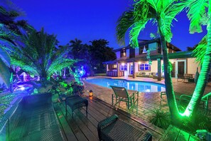 A swimming pool with a wooden deck, lounge chairs, and a house with illuminated windows. - Blockchain Manor (North Miami Beach)