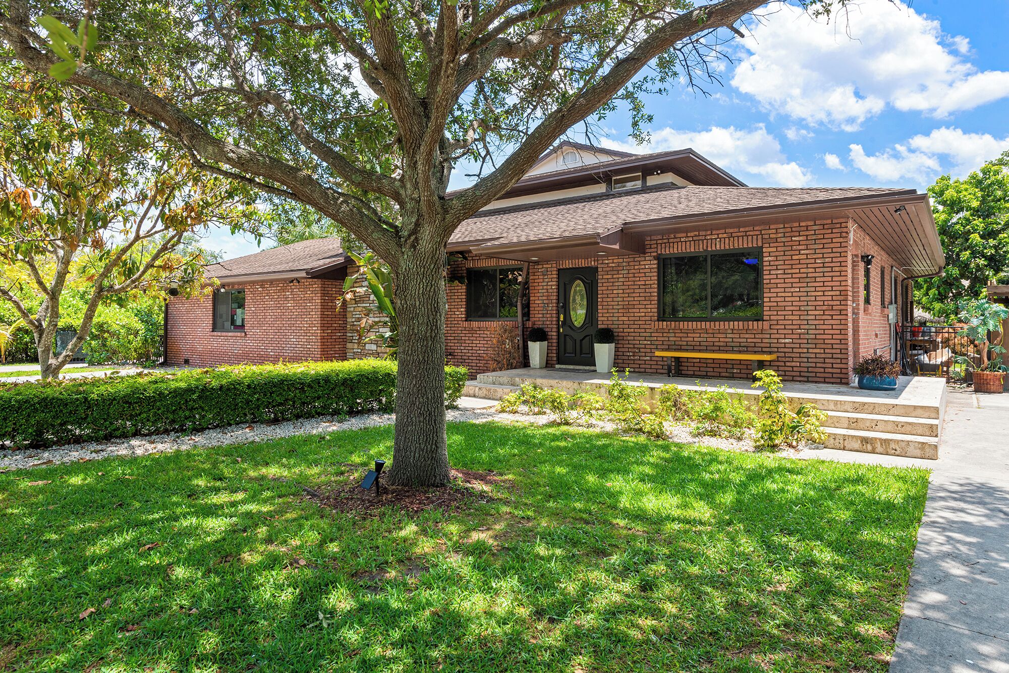 A brick house with a front porch, a tree in front, and a bench.