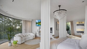 A modern bedroom with a curved ceiling, a large bed, a seating area, and a view of trees through large windows. - Beverly Crest (Beverly Hills)