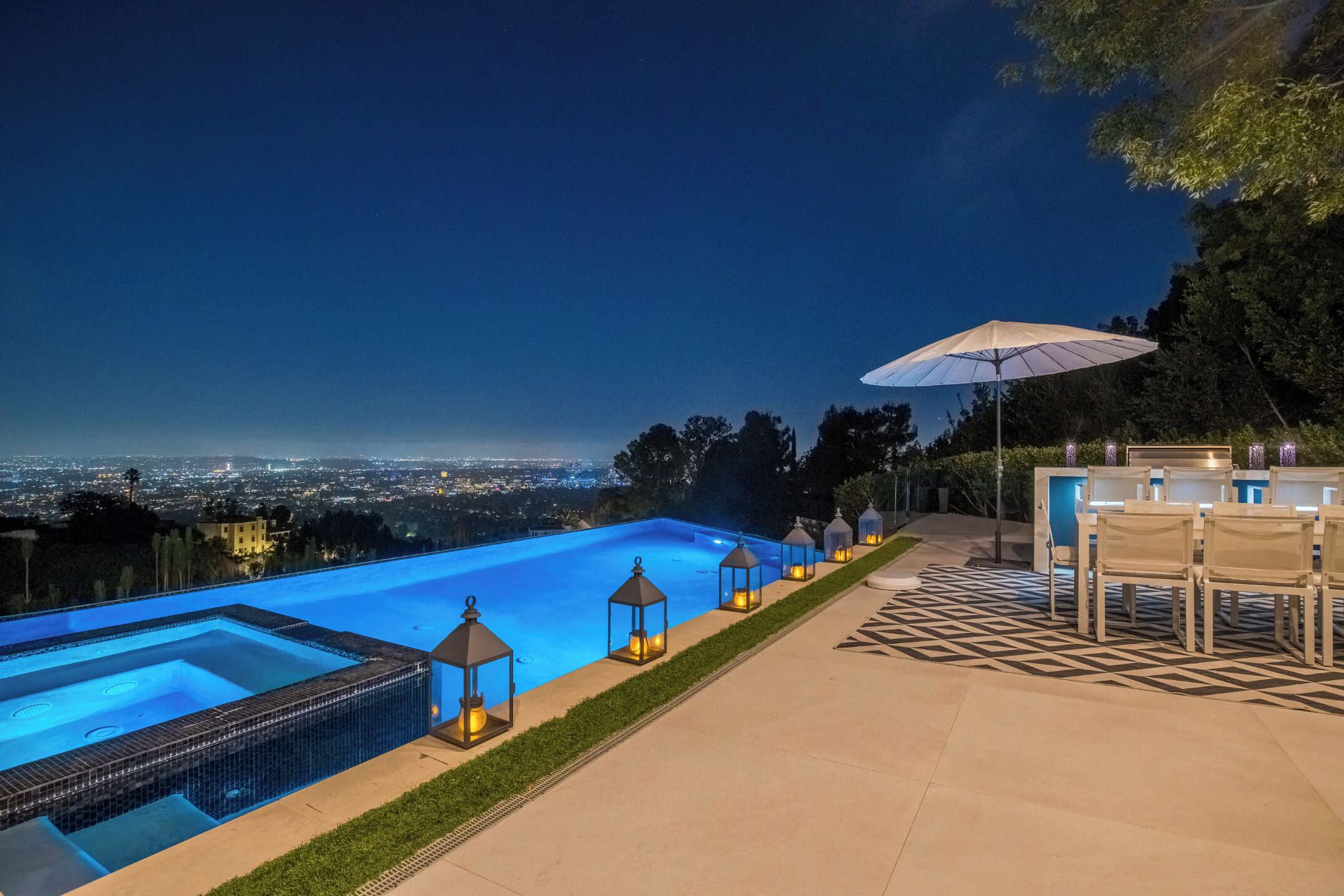 Una piscina con vistas a un paisaje urbano nocturno, rodeada de mobiliario exterior y faroles.