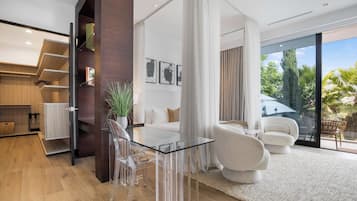A modern living room with a glass table, clear chairs, a white sofa, and a large window with a view of greenery.