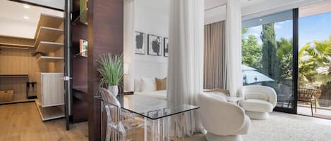 A modern living room with a glass table, clear chairs, a white sofa, and a large window with a view of greenery.