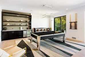 A modern living room with a pool table, a sofa, and a large window offering a view of greenery. - Saint Ives Reserve (West Hollywood)