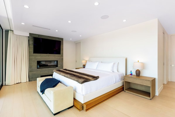 Una habitación de hotel moderna con una cama grande, un televisor de pantalla plana montado en una pared de piedra, una mesita de noche con una lámpara y una pequeña zona de estar con una silla y una manta.