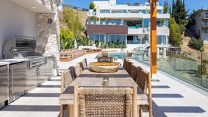 Un área de comedor al aire libre con una parrilla, asientos y vistas a una piscina y edificios.