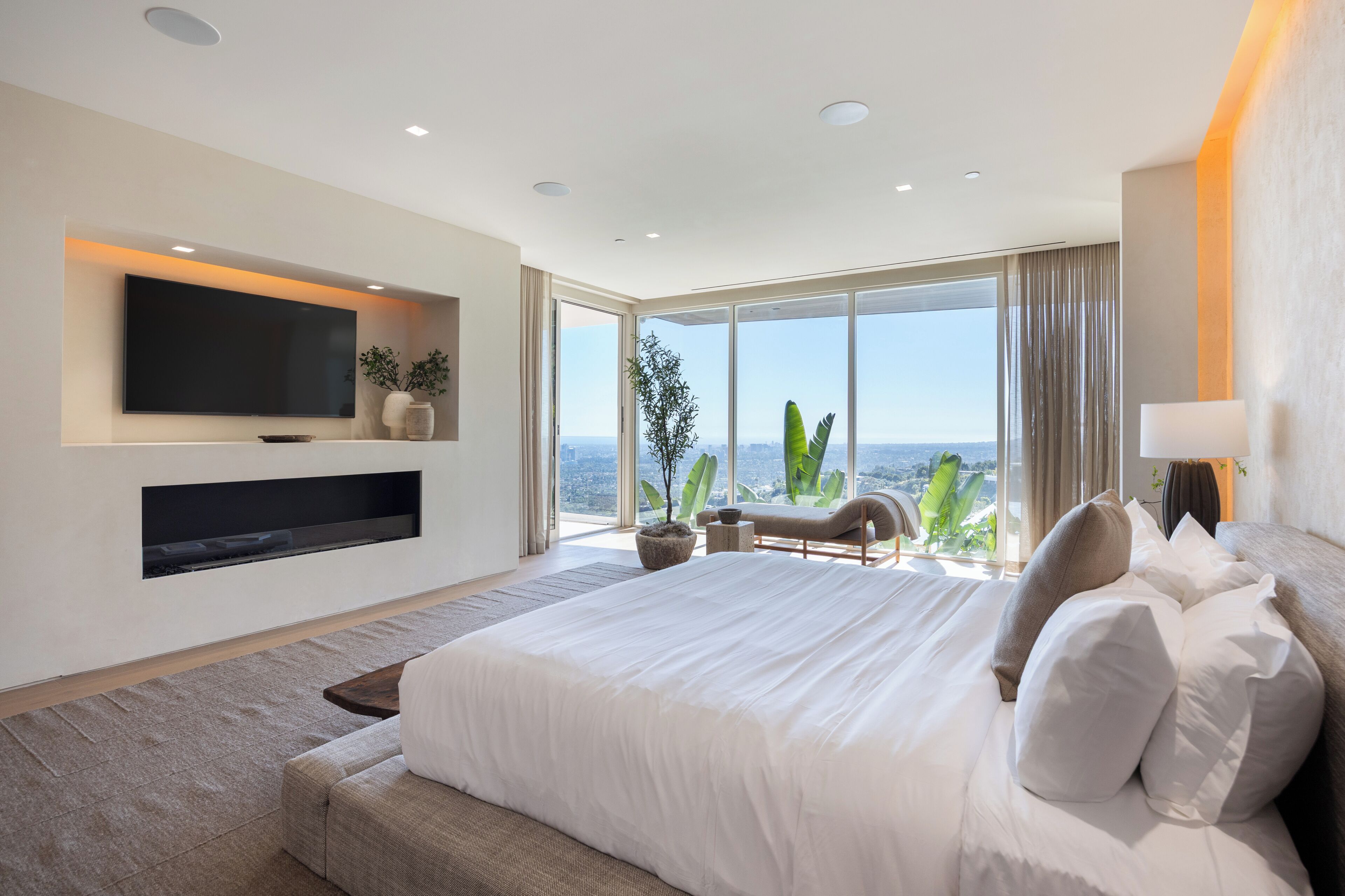 A modern bedroom with a large bed, a mounted TV, a view of the city, and a balcony with plants.