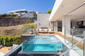 Un área de piscina moderna al aire libre con un muro de piedra, una terraza de madera y un balcón con cerramiento de vidrio.