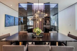 Ein moderner Essbereich mit einem dunklen Holztisch, gepolsterten Stühlen, einer geometrischen Leuchte und einem Weinkühlschrank im Hintergrund.