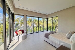 Ein modernes Schlafzimmer mit einem großen Bett, einer Couch und einem Balkon mit Blick auf Palmen.