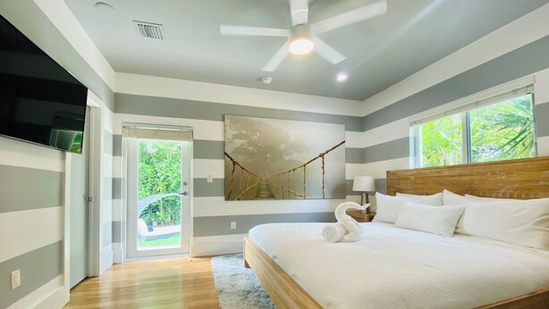 Une chambre avec un lit en bois, un ventilateur de plafond, une grande fenêtre donnant sur un espace verdoyant et une porte menant à un espace extérieur.