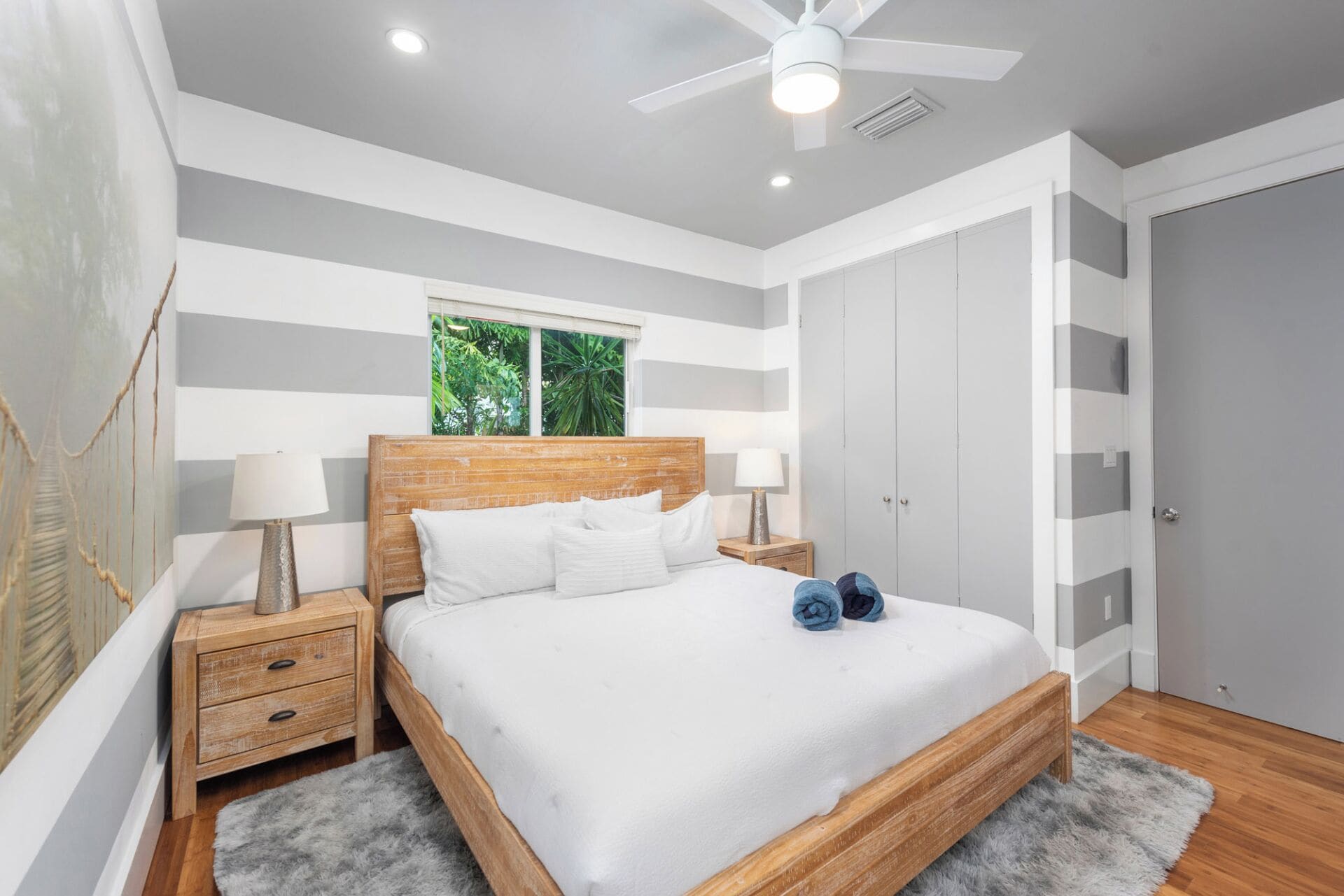 A bedroom with a bed, bedside tables, a ceiling fan, and a window with a view of greenery.