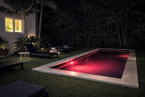 A swimming pool with a red light, surrounded by lounge chairs and potted plants, with a building in the background.