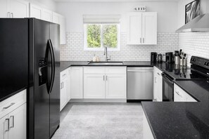 A modern kitchen with white cabinets, a black refrigerator, a window with a view of greenery, and a black countertop.