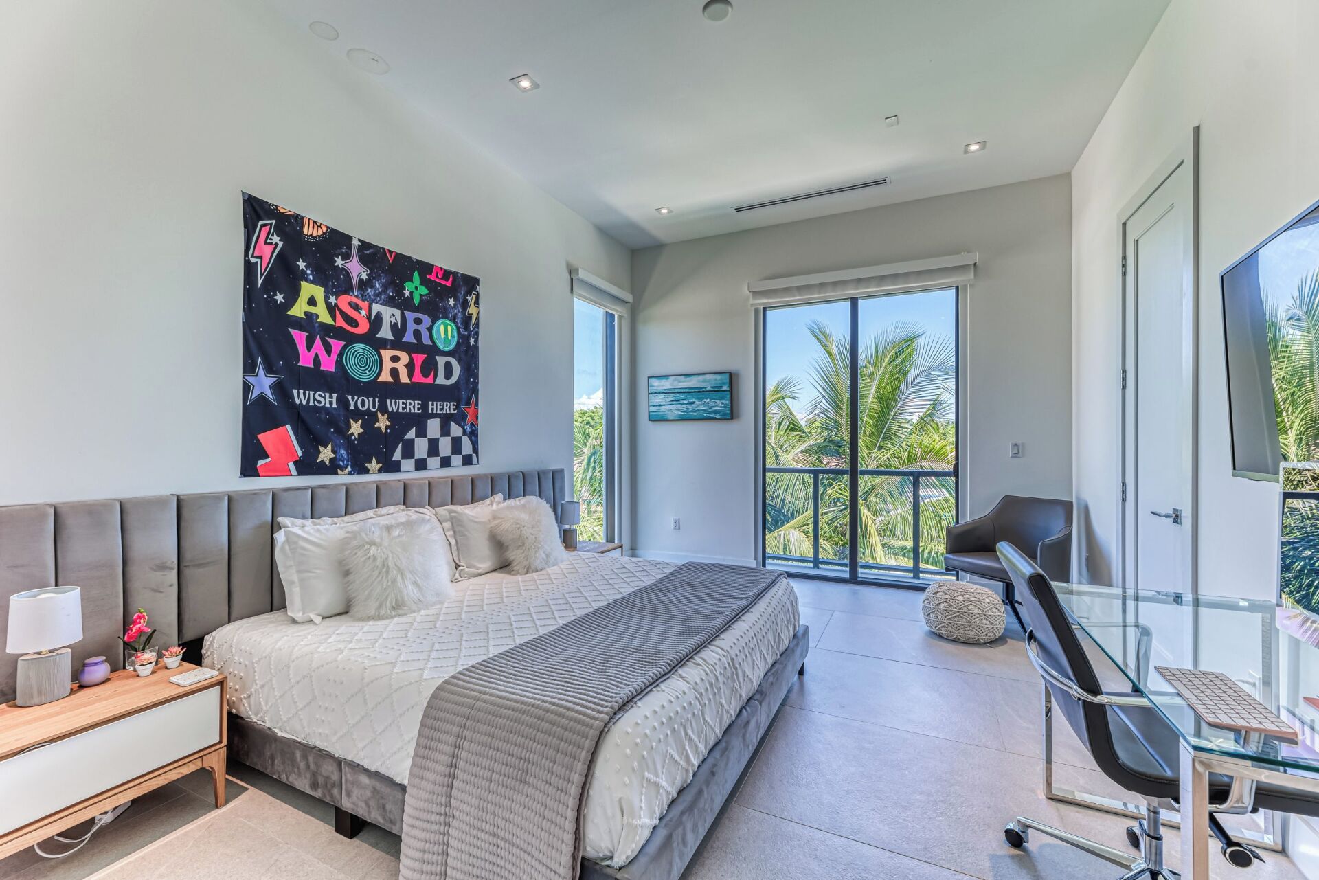A modern bedroom with a large bed, a desk, and a view of palm trees.