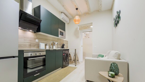 Et kompakt køkken med moderne hårde hvidevarer, et spiseområde med et lille bord, og en opholdsstue med en sofa.